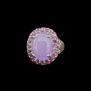 May include: A silver ring featuring a large, oval, light purple gemstone surrounded by smaller, round, pink gemstones. The ring is set against a black background, highlighting the colors and design of the jewelry.