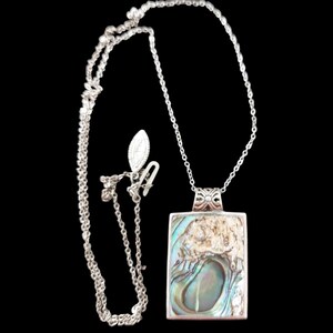 Op de afbeelding: Een zilveren ketting met een rechthoekige hanger met een abalone schelp ontwerp. De hanger is omlijst in zilver en hangt aan een delicate ketting. De achtergrond is zwart.