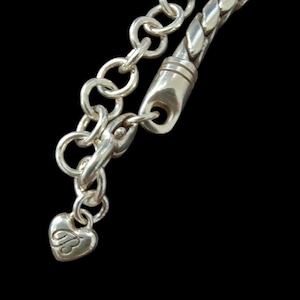 Puede incluir: Primer plano de un collar plateado con un dije en forma de coraz&oacute;n. El collar presenta un dise&ntilde;o de eslabones de cadena y una secci&oacute;n trenzada. El dije de coraz&oacute;n tiene una letra "B" estilizada grabada. El fondo es negro.