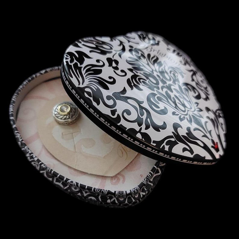 May include: Heart-shaped trinket box with a black and white damask pattern. The box is open, revealing a small silver charm with gold accents. The interior has a pink heart-shaped card. The box is on a black background.