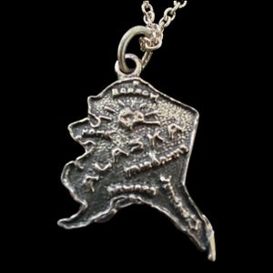 Op de afbeelding: Een zilverkleurige ketting met hanger met een gedetailleerde omtrek van de staat Alaska. De hanger heeft een getextureerd oppervlak met gegraveerde details, waaronder het woord "Barrow" bovenaan. De ketting hangt aan een fijne zilveren ketting.