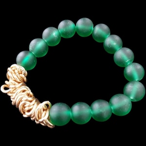 May include: A bracelet featuring large, translucent green beads strung together. The bracelet is accented with a gold-colored, intricately woven clasp. The beads have a frosted appearance, and the background is black.