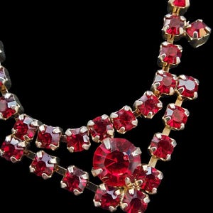 Puede incluir: Primer plano de un collar vintage con engaste dorado y vibrantes piedras rojas. El collar presenta un diseño en capas con piedras cuadradas y redondas, creando un accesorio brillante y elegante.