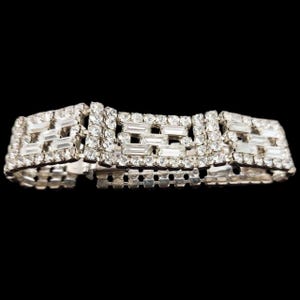 May include: A silver-toned bracelet with rectangular and round clear rhinestones. The bracelet has three rectangular sections with a geometric design, connected by a chain of small, round rhinestones. The background is black.