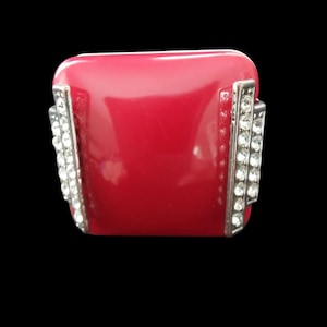 May include: A striking, red rectangular ring with a glossy finish. The ring has silver-toned accents on the sides, adorned with rows of sparkling, clear crystals. The design is reminiscent of Art Deco jewellery.