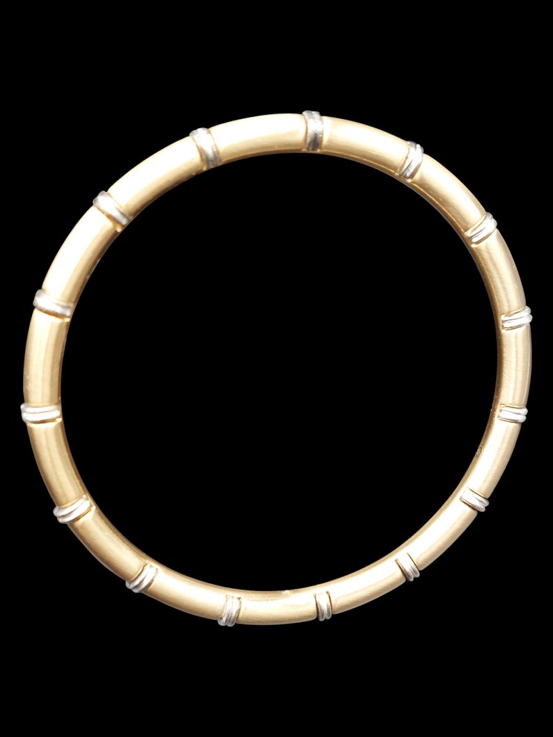 K&ouml;nnte beinhalten: Ein goldfarbenes Armreif-Armband mit Bambus-Design. Das Armband hat Segmente, die durch silberfarbene B&auml;nder getrennt sind. Das Armband ist kreisf&ouml;rmig und vor einem schwarzen Hintergrund platziert.