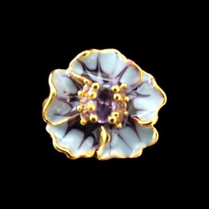 May include: A flower-shaped brooch featuring light blue petals with dark purple veining and gold-toned edges. The centre of the flower is adorned with small gold beads and a central purple gemstone, creating a detailed floral design.