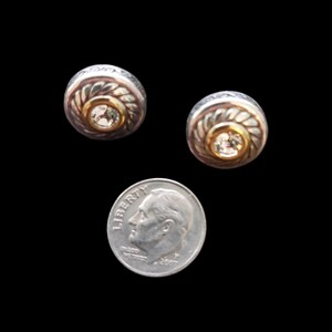 May include: Two silver-toned stud earrings with a gold-toned center and a clear gemstone. The earrings have a textured, circular design. A U.S. dime is included for size comparison. The background is black.