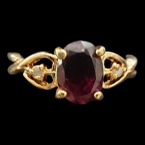 May include: A gold ring with an oval, deep red gemstone as the centerpiece. The ring has a delicate, ornate design with small accent stones on either side of the main stone. The background is black.