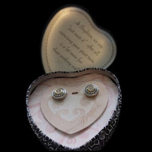 May include: A pair of silver and gold stud earrings with a clear stone center, displayed in a heart-shaped box. The box is black and white patterned with a heart-shaped interior. The box lid has text that reads "At Brighton, we say just wear it..."