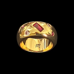 May include: A wide gold band ring featuring a polished finish with an irregular surface. The ring is set with one rectangular red gemstone and several smaller round pink and red gemstones. The interior of the band is engraved with the letter m.