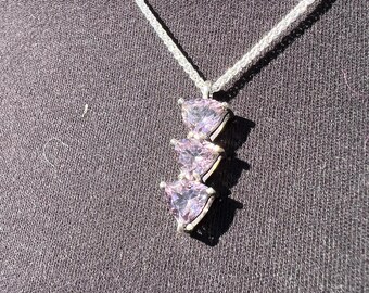May include: A silver necklace with a pendant featuring three heart-shaped, light purple gemstones. The chain is a delicate, woven design. The pendant is centered against a dark grey background.