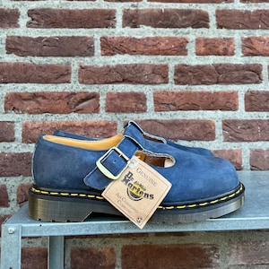 May include: Navy blue Dr. Martens Mary Jane shoes with a buckle closure and yellow stitching. The shoes have a thick, black sole and a tag that reads "Genuine Dr. Martens Air Cushioned Comfort."