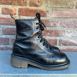 May include: Black leather ankle boots with a lace-up design. The boots have a rounded toe, a thick sole, and a small heel. The brand name is visible on the side of the boot. The boots are set against a brick wall.