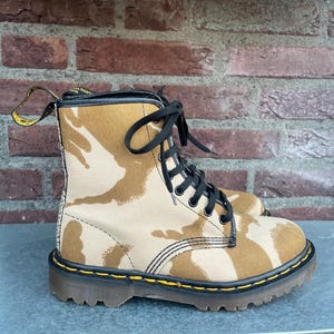 May include: Tan and brown camouflage print combat boots with black laces and a yellow pull tab. The boots have a black sole with yellow stitching and a brown rubber outsole. The boots are made of canvas and leather.