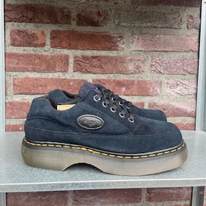 Può includere: Sneakers platform in camoscio nero con punta arrotondata e suola spessa cucita in giallo. Le sneakers hanno lacci neri e un piccolo logo ovale. Altezza di circa 5 cm.