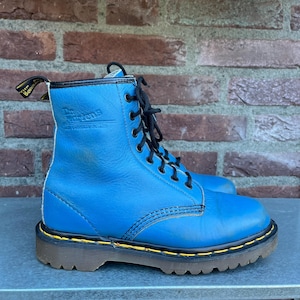 May include: A pair of blue leather boots with black laces. The boots have a yellow trim around the sole and a brown rubber sole. The brand name "Dr. Martens" is visible on the side of the boot.