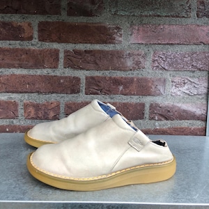 May include: A pair of light beige slip-on clogs with a thick, tan sole. The clogs have a rounded toe and a smooth leather-like upper. The background features a brick wall.