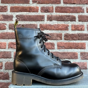 May include: Black leather combat boot with a lace-up design. The boot features a sturdy sole and a pull tab with yellow text. The boot is set against a brick wall background.