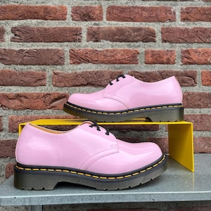 Może przedstawiać: Dwie pary różowych lakierowanych butów ze sznurowadłami i czarnymi podeszwami. Buty mają żółte szwy wokół podeszew. Buty są wystawione na żółtej półce przed ceglaną ścianą.