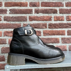 Op de afbeelding: Een zwarte leren enkelboot met een zijband en een zilverkleurige metalen gesp waarop de tekst DM's is gegraveerd. De schoen heeft een ronde neus, een zichtbare naad aan de voorzijde en een doorschijnende, geribbelde rubberen blokhak en zool.
