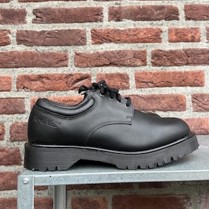 May include: Black leather shoe with a thick, lugged sole. The shoe has a rounded toe and a lace-up closure. The words "Royal Mail" are embossed on the side of the shoe. The shoe is sitting on a metal surface in front of a brick wall.