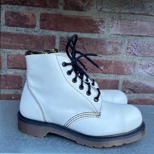 May include: White leather ankle boots with black laces and stitching. The boots have a brown sole and a classic design. The boots are on a gray surface with a brick background.