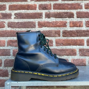 May include: A pair of navy blue leather boots with green laces and yellow stitching around the soles. The boots are sitting on a metal surface in front of a brick wall.