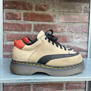 May include: Tan and black platform sneakers with black laces and a red heel accent. The shoes have a thick, brown sole with yellow stitching. The brand name is visible on the side of the shoe.