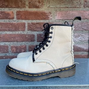 May include: Tan canvas lace-up boots with black laces and a black trim. The boots have a yellow stitched sole and a pull tab at the back. The boots are set against a brick wall background.