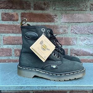 May include: Black canvas Dr. Martens boots with a white stitched sole. The boots have a lace-up design, metal eyelets, and a branded tag. The boots are displayed on a gray surface against a brick wall.