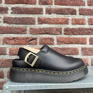 Pode incluir: Um sapato tipo soca em couro preto com sola de plataforma grossa e costuras amarelas. O sapato apresenta uma tira traseira ajustável com uma fivela de metal dourado gravada com o texto Dr. Martens e uma biqueira arredondada.