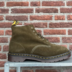 Peut inclure: Une bottine en daim vert olive à lacets, dotée d'œillets métalliques dorés et d'une épaisse semelle en caoutchouc marron foncé avec des surpiqûres jaunes. Une tirette en tissu jaune et noir est fixée à l'arrière du col de la chaussure.