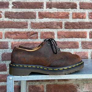 May include: Brown leather Oxford-style shoes with black laces. The shoes have a yellow stitch detail around the sole and a dark brown sole. The shoes are sitting on a metal surface in front of a brick wall.