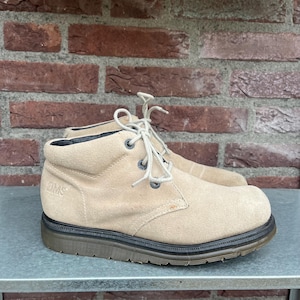 May include: A pair of tan suede ankle boots with light-colored laces and a dark brown sole. The boots have a rounded toe and a low heel. The word "IMS" is visible on the side of one boot. The boots are sitting on a metal surface.