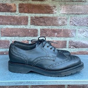 May include: Black leather brogue shoes with a lace-up closure. The shoes have decorative perforations and a thick, lugged sole. The shoes are displayed on a blue surface against a brick wall.