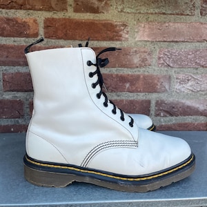 May include: A pair of white leather boots with black laces. The boots have a classic design with a yellow trim around the sole and black stitching. The boots are approximately 10 inches tall.