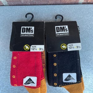May include: Two pairs of Dr. Martens socks, one red and one black, each with a gold-colored toe and heel. The socks have small, gold-colored dots and the text "LYCRA" on a white label. The packaging reads "DM's DR. MARTENS".