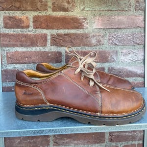 Op de afbeelding: Bruine leren veterschoenen met zichtbare stiksels en een dikke, donkere zool. De schoenen hebben een klassiek ontwerp, geschikt voor vrijetijdskleding. De merknaam is zichtbaar aan de zijkant.