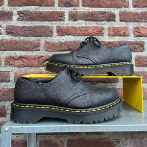 Peut inclure: Une paire de chaussures à plateforme noires à paillettes avec des lacets noirs et des coutures jaunes. Les chaussures sont exposées sur une étagère jaune contre un mur de briques.