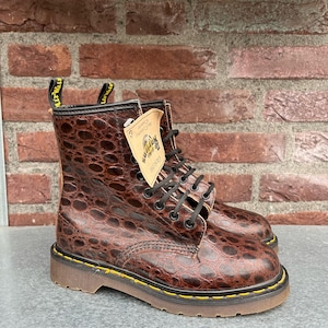 Könnte beinhalten: Braune Leder-Ankle Boots mit Krokodilmuster. Die Stiefel haben schwarze Schnürsenkel, gelbe Nähte und eine braune Sohle. Ein Etikett ist angebracht. Die Stiefel werden vor einer Backsteinmauer präsentiert.