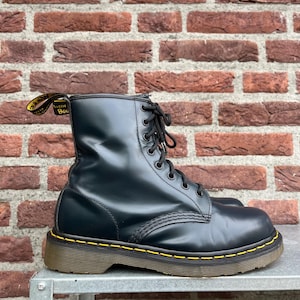 Puede incluir: Bota de combate de cuero negro con diseño de cordones y costuras amarillas alrededor de la suela. La bota tiene una lengüeta con texto amarillo. La bota está sobre una superficie metálica frente a una pared de ladrillos.