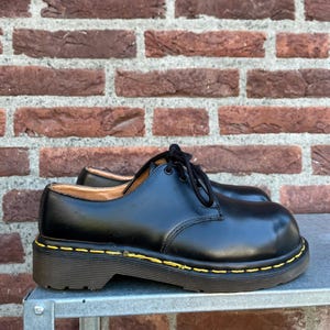 May include: Black leather shoes with a low heel and yellow stitching. The shoes have a classic lace-up design and a rounded toe. The shoes are sitting on a metal surface.