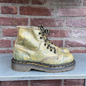 May include: A pair of olive green and yellow leather ankle boots with black and yellow laces. The boots have a thick, dark brown sole with yellow stitching and a pull tab at the top.