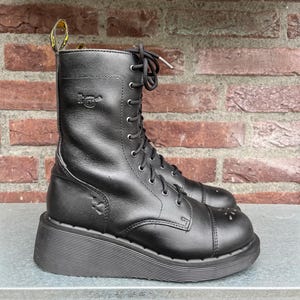 May include: Black leather combat boots with a platform sole. The boots have a lace-up design, a rounded toe, and a pull tab at the top. The boots are set against a brick wall background.