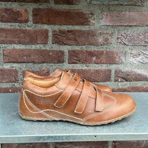 Può includere: Scarpe in pelle marrone con tre cinturini regolabili. Le scarpe hanno la punta arrotondata e sono cucite con filo chiaro. Le suole sono marrone chiaro. Le scarpe misurano circa 25 cm di lunghezza.