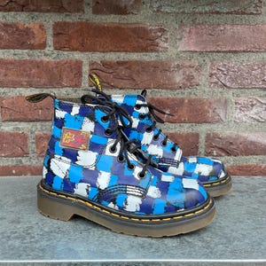 May include: A pair of blue, white, and black patterned boots with black laces. The boots have a yellow stitched sole and a small patch with a floral design. The boots are on a gray surface with a brick background.
