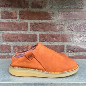 May include: Orange slip-on shoe with a rounded toe and a light tan sole. The shoe has a stitched design around the sole and a small label that reads "DMS". The shoe is made of a suede-like material.