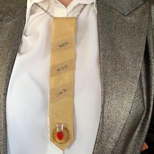 May include: A tan-colored, rectangular pendant necklace with a chain. The pendant has a striped pattern with small, decorative embellishments. The necklace is worn over a white collared shirt and a metallic gold blazer.