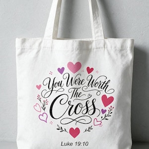 Könnte beinhalten: Eine weiße Tragetasche aus Segeltuch mit dem schwarzen kursiven Schriftzug You Were Worth The Cross und dem Verweis Luke 19:10 darunter. Das Design ist von verschiedenen rosa, roten und violetten Herzillustrationen und kleinen schwarzen Blumenranken umgeben.